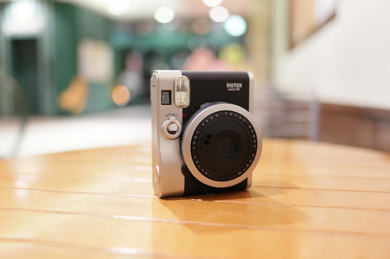 Black and silver Fujifilm instax mini 90 camera on a wooden table, with mini 90 marking visible