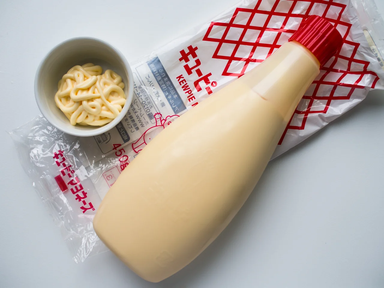 Japanese Kewpie standard mayonnaise 450g bottle with red cap and red-grid outer bag, beside a bowl of mayonnaise; older 2016 packaging